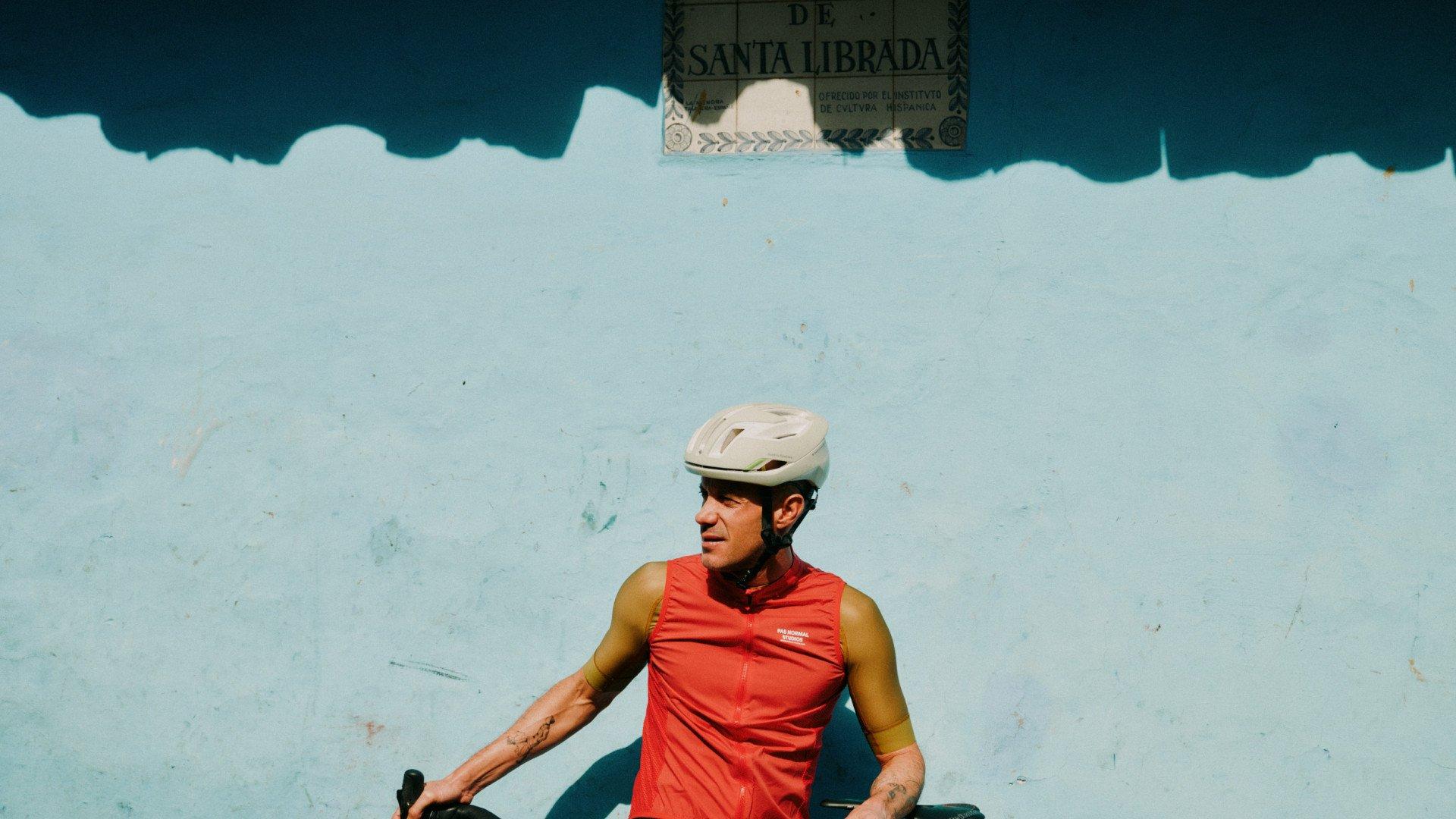Model wearing red sleeveless cycling jersey with white helmet against blue wall