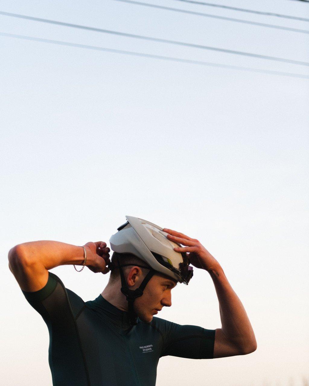 Model wearing dark green cycling jersey adjusting white helmet against cloudy sky