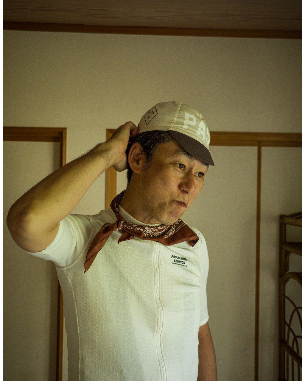Model wearing white jersey with chest logo and brown bandana, tan cap with white lettering indoors