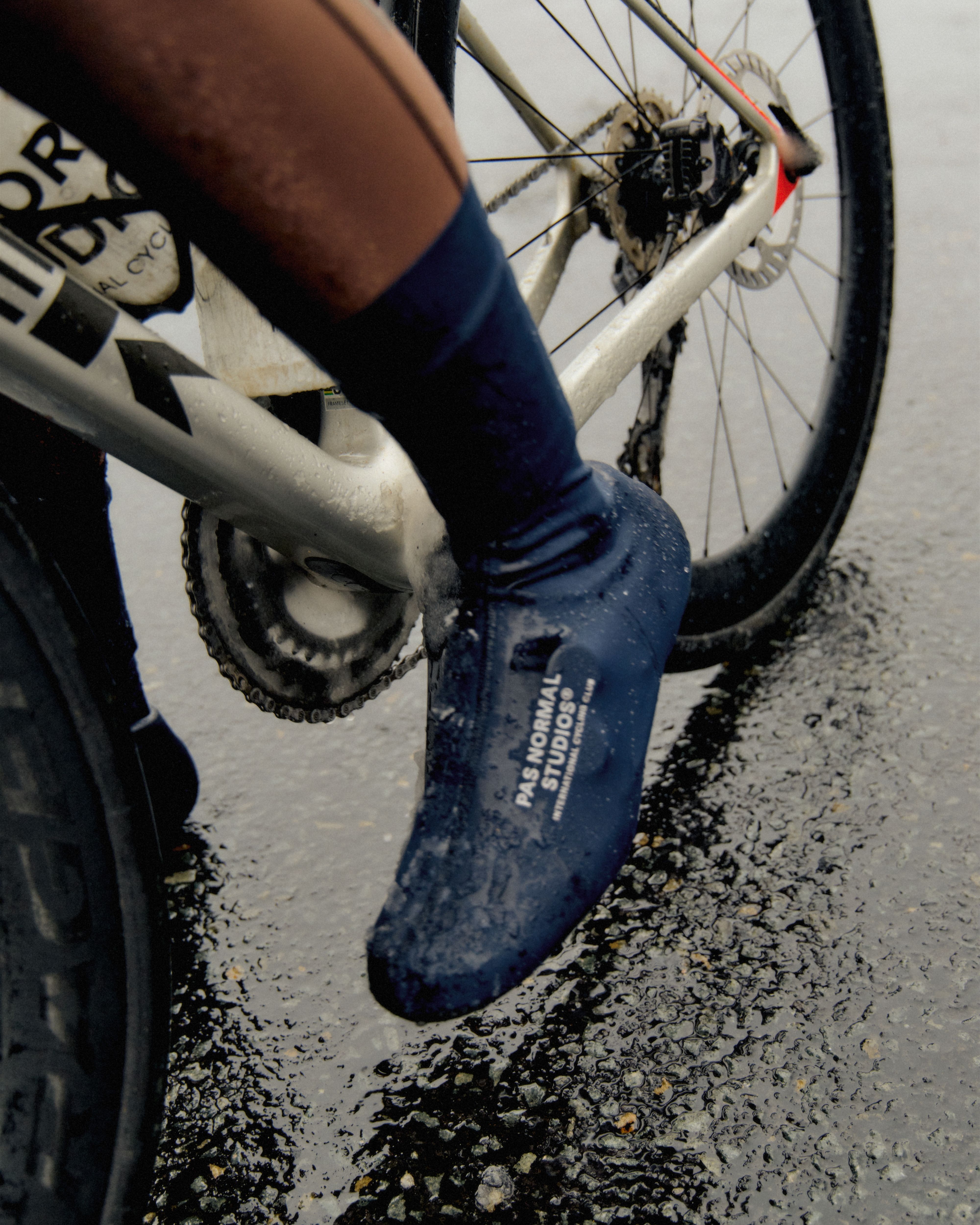Model wearing blue overshoes with white branding, standing next to white bicycle on wet road