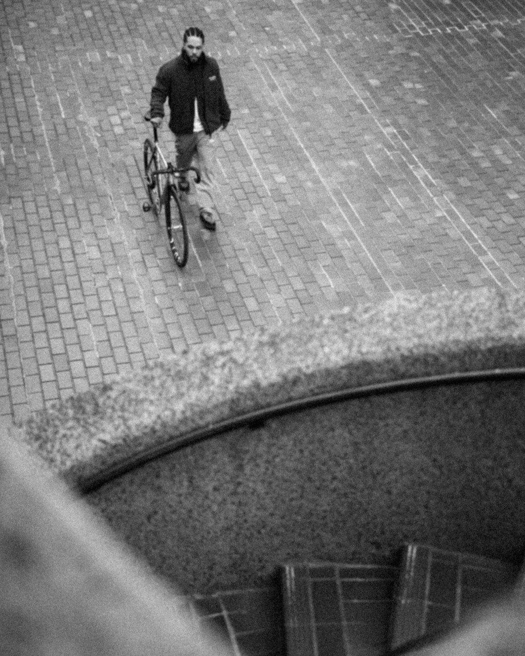 Model wearing dark jacket walking with bicycle on cobblestone street, black and white street photography