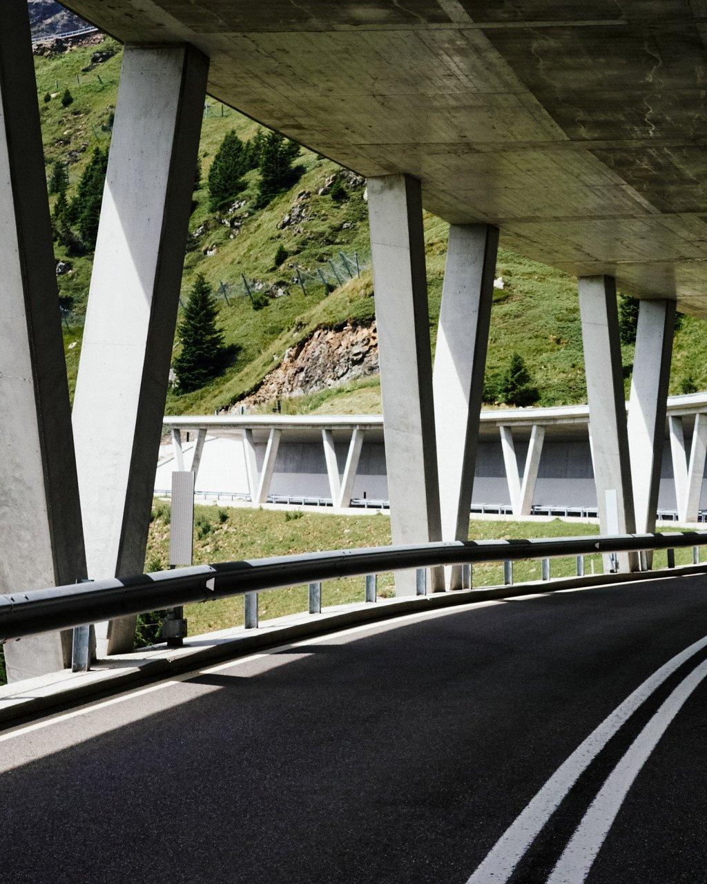 Modern concrete bridge with angular supports over mountain road with green alpine hills