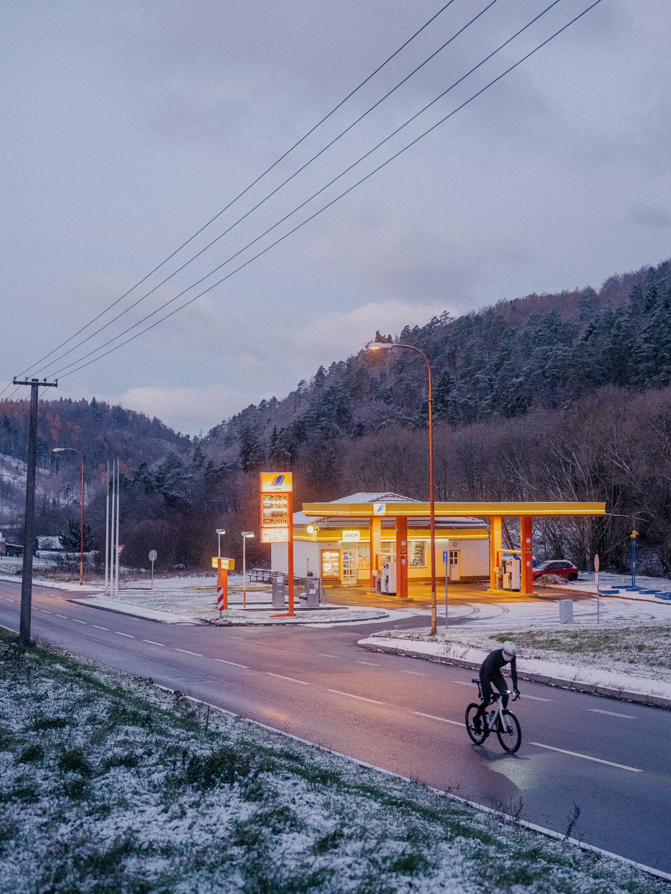 Cyclist riding past illuminated gas station at dusk in snowy mountain landscape