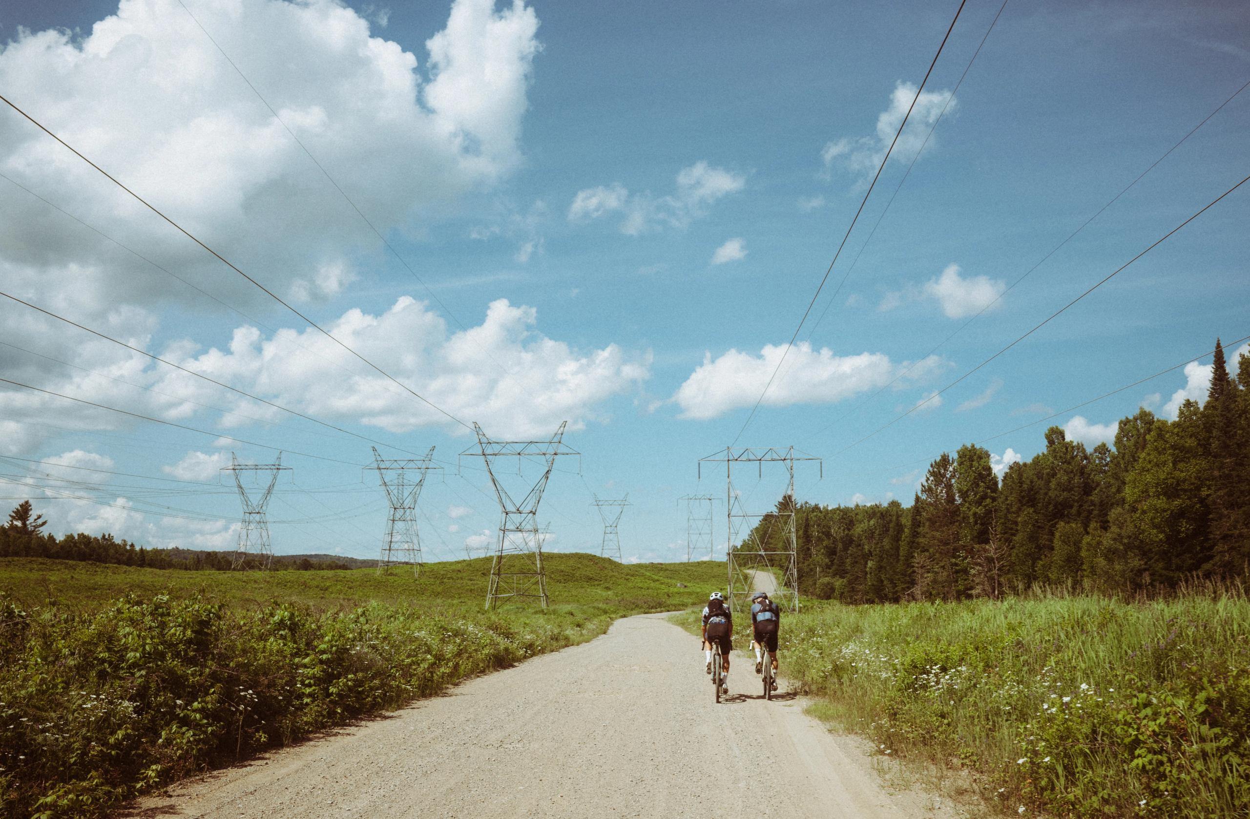 Two cyclists riding on rural road through countryside with power lines overhead