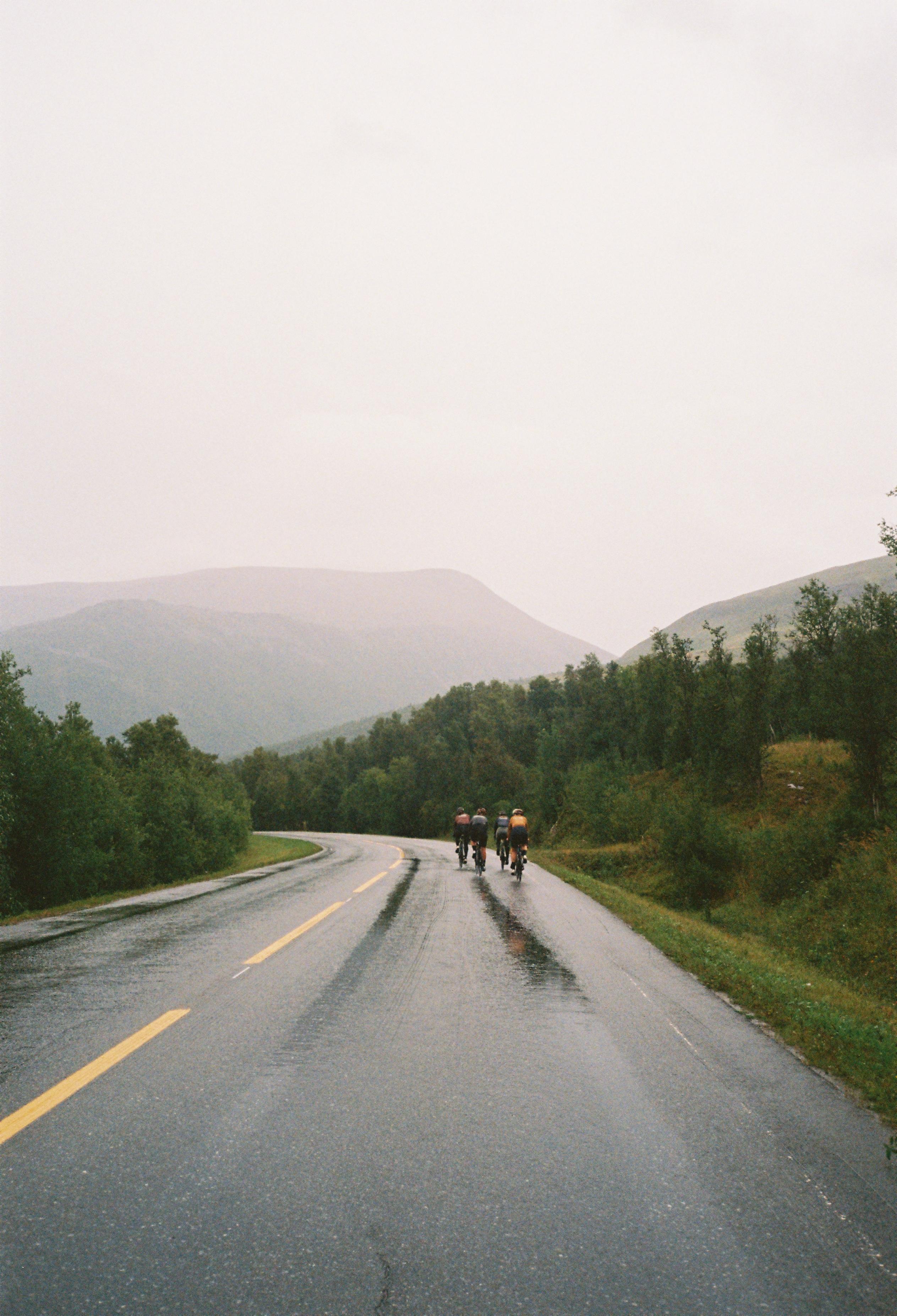 Cyclists riding on wet mountain road through forested hills in misty conditions