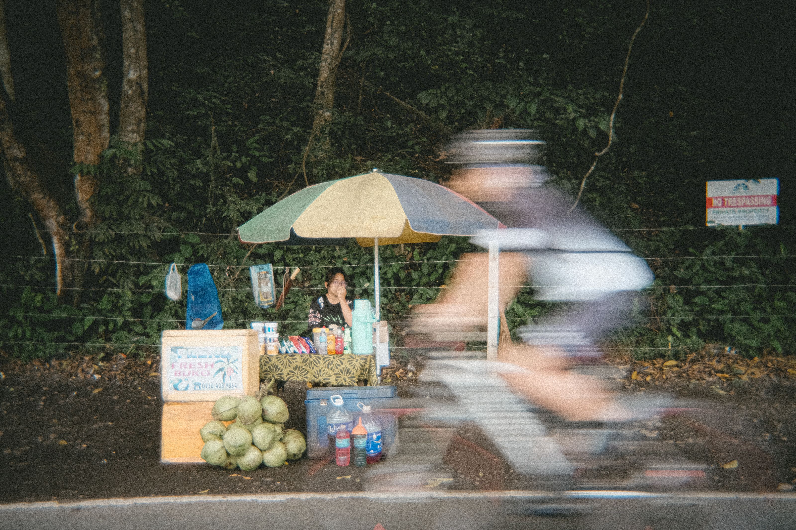 Motion-blurred cyclist passing roadside fruit stand with vendor under umbrella