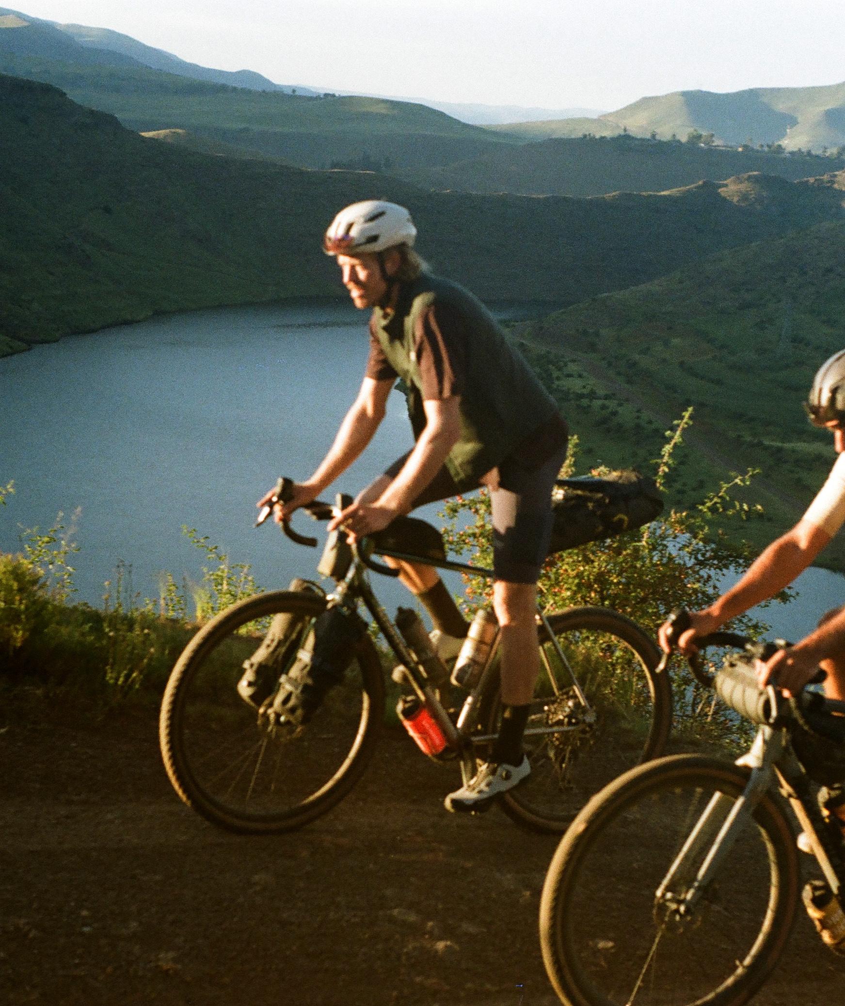 Two cyclists riding mountain bikes on hillside trail overlooking lake and mountains in golden light