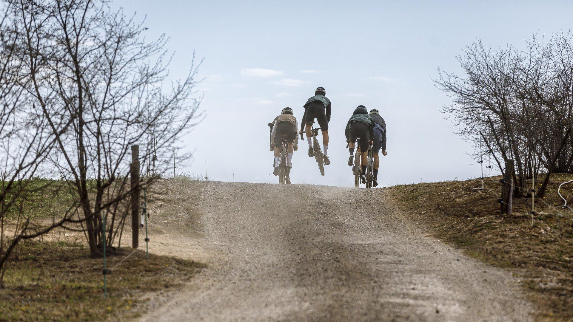 Four cyclists riding gravel bikes up dusty hill with bare trees under overcast sky