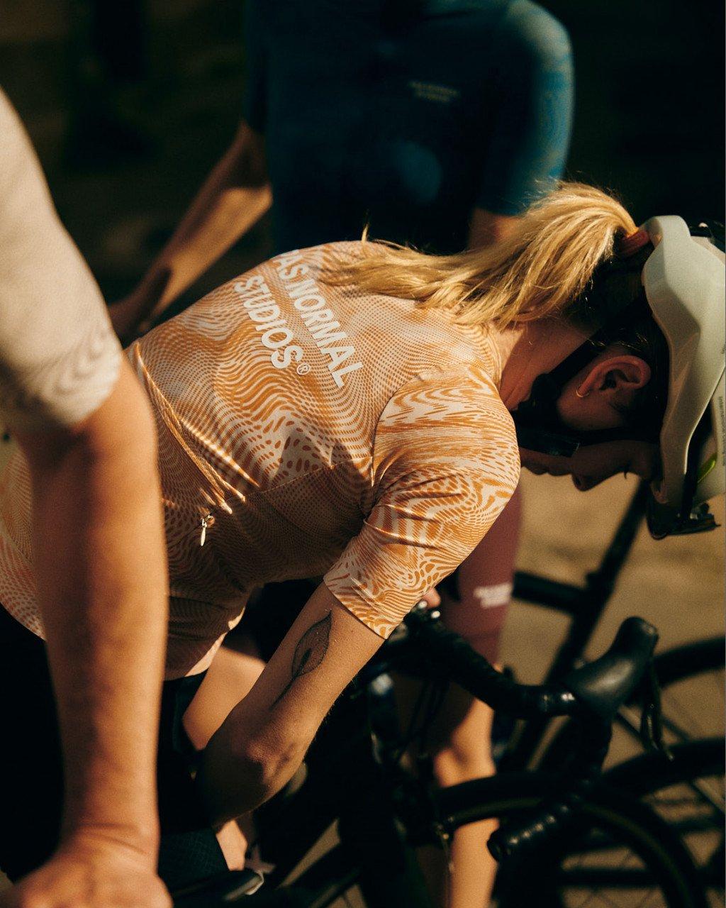 Model wearing orange patterned cycling jersey with back lettering while riding indoor trainer