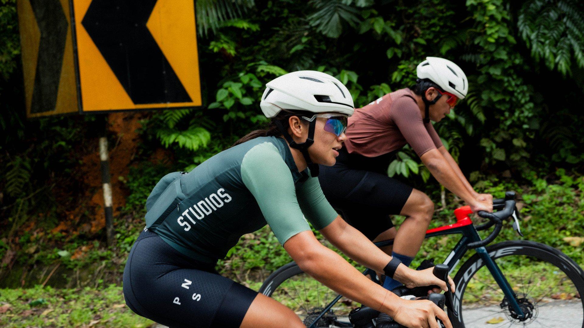 Models wearing cycling apparel in aerodynamic position, woman in green and navy bodysuit with branding on back