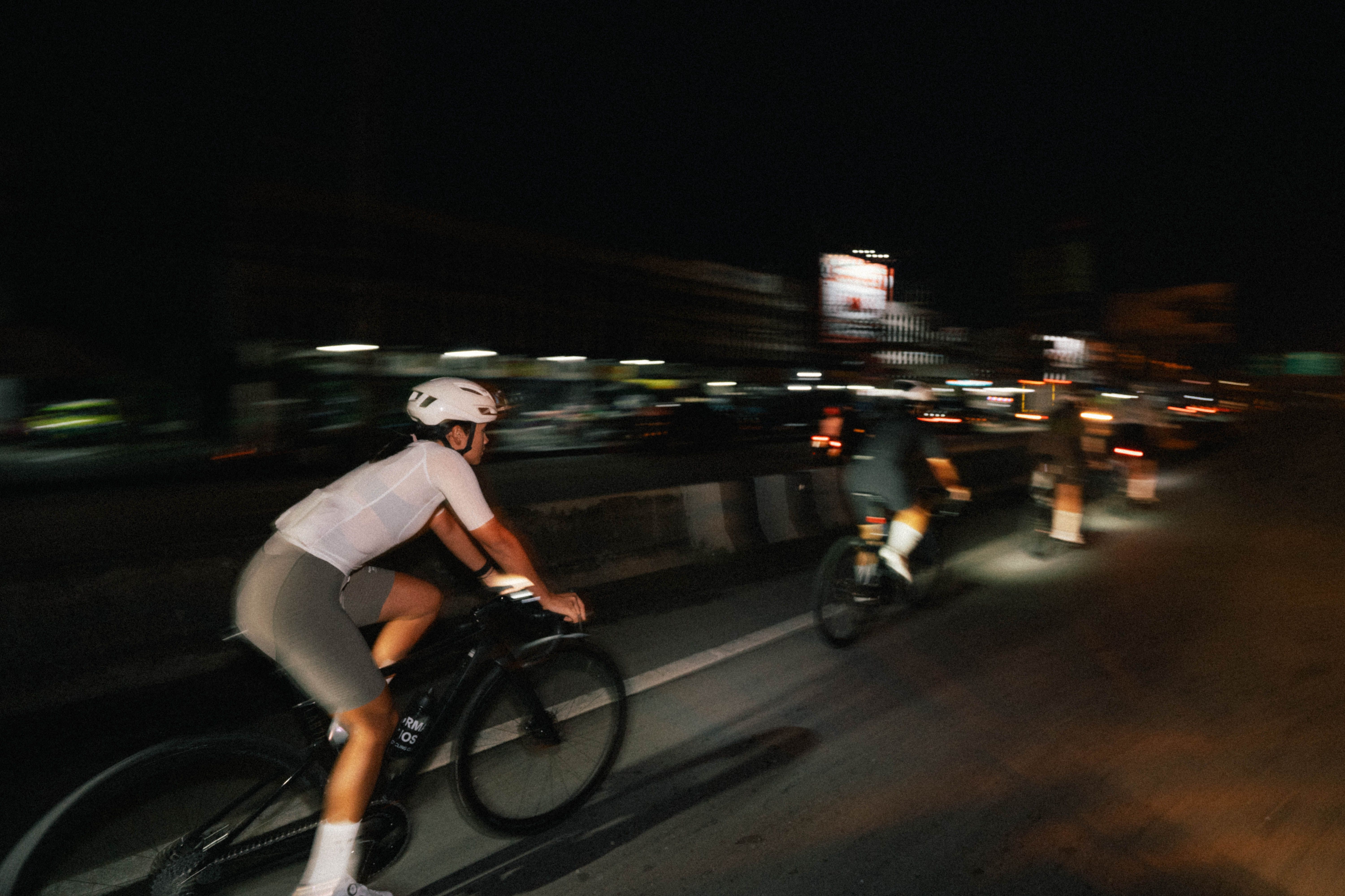 Model wearing white jersey and grey shorts cycling at night through urban streets with motion blur