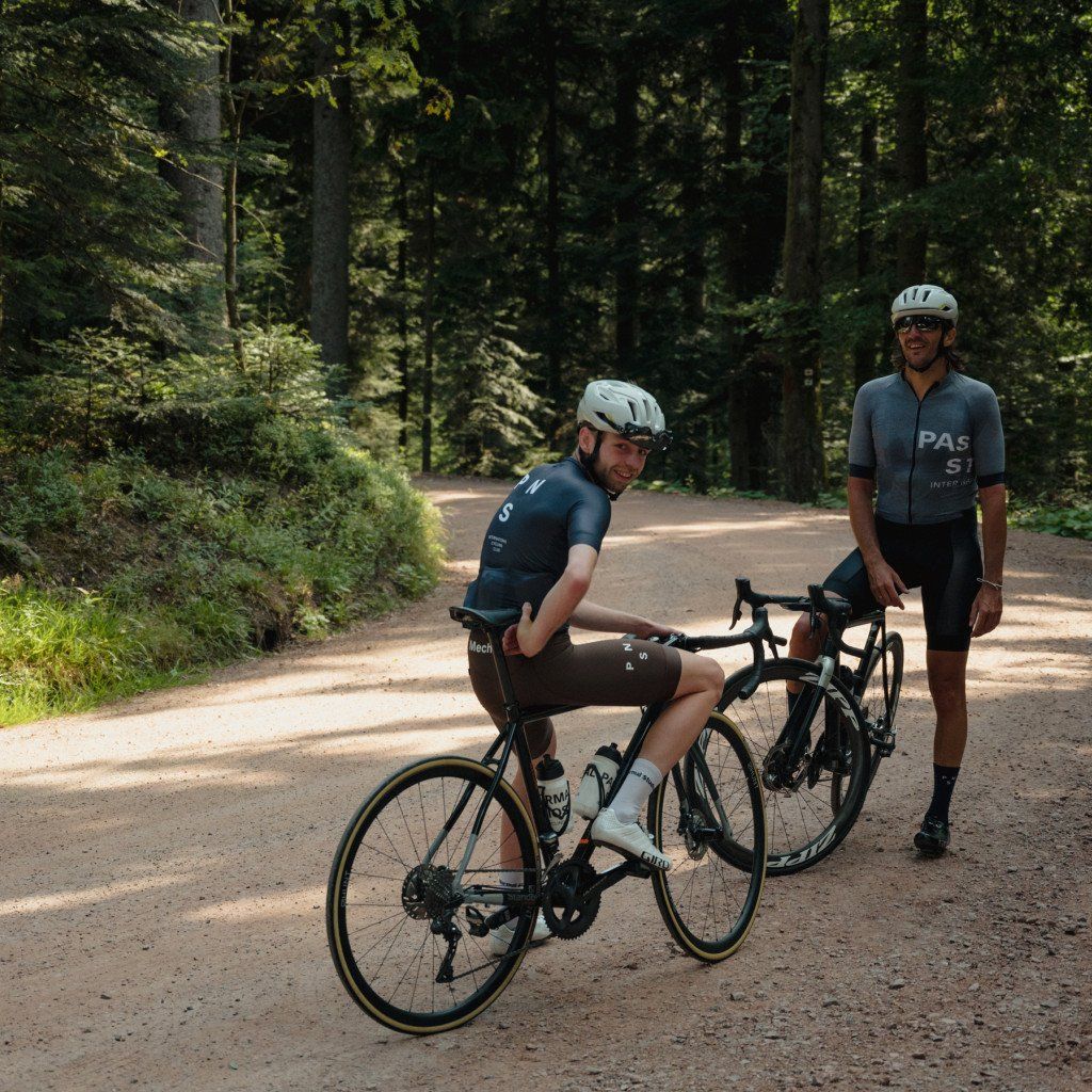 Two cyclists wearing PAS collection jerseys with one on bike and one standing on forest road