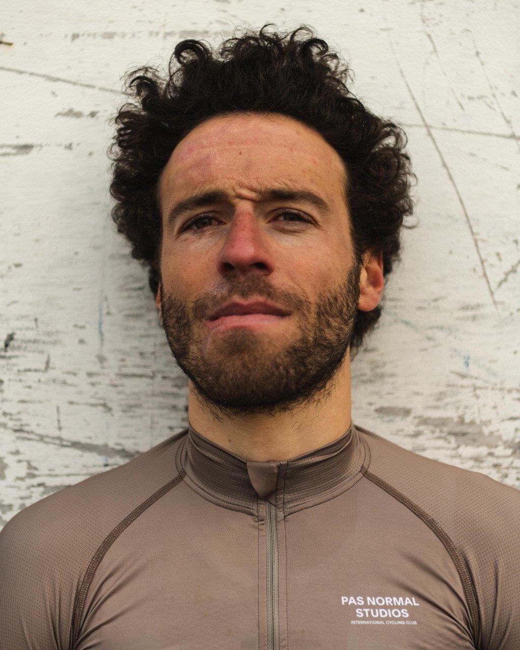 Model wearing brown zip-up cycling jersey with white branding against white brick wall