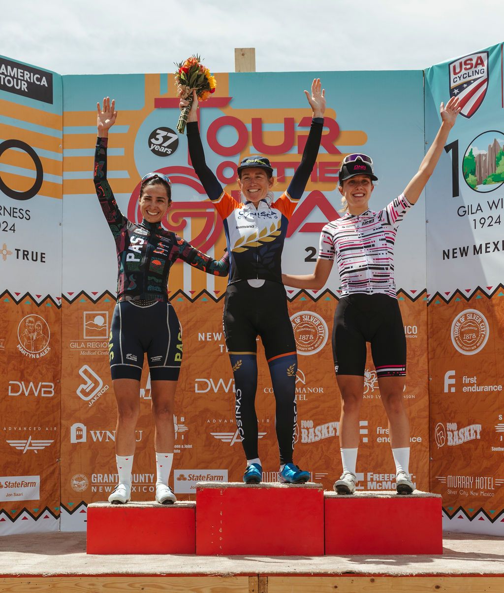 Three female cyclists on podium celebrating race victory, wearing professional racing jerseys and raising arms in triumph