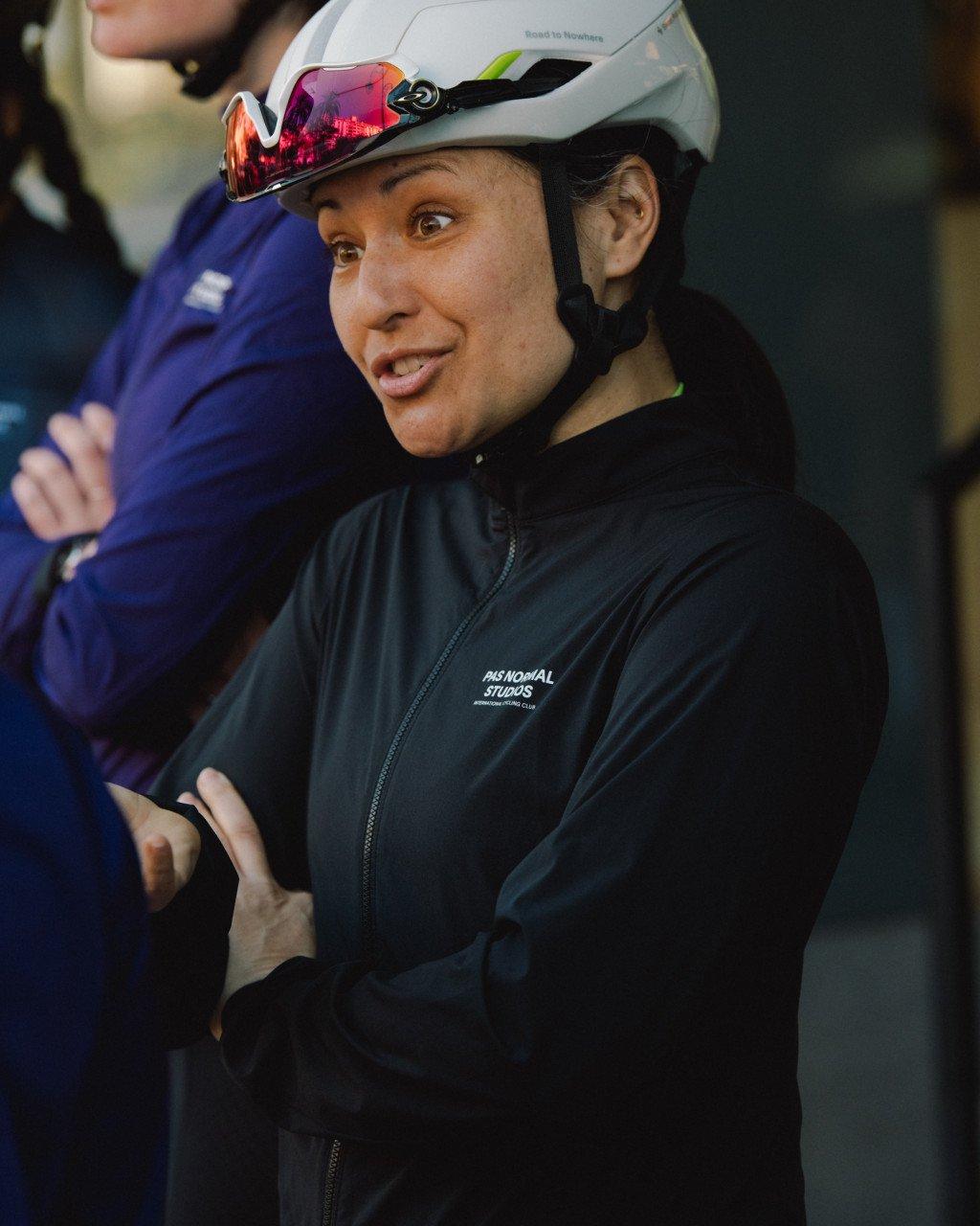 Model wearing black zip-up jacket with white aero helmet, smiling in cycling environment