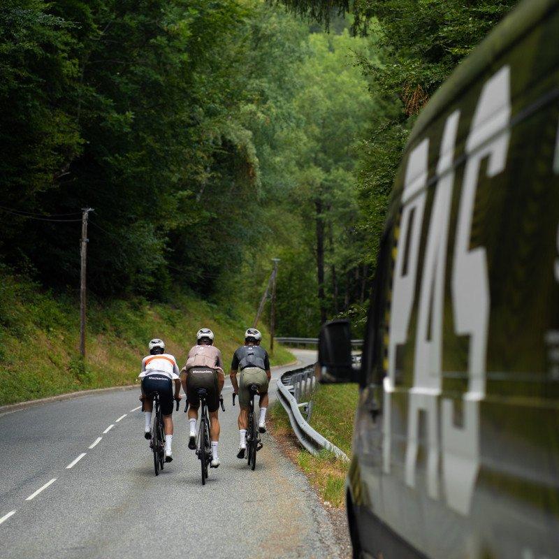 Three cyclists in aerodynamic position on rural road with support vehicle, surrounded by green forest