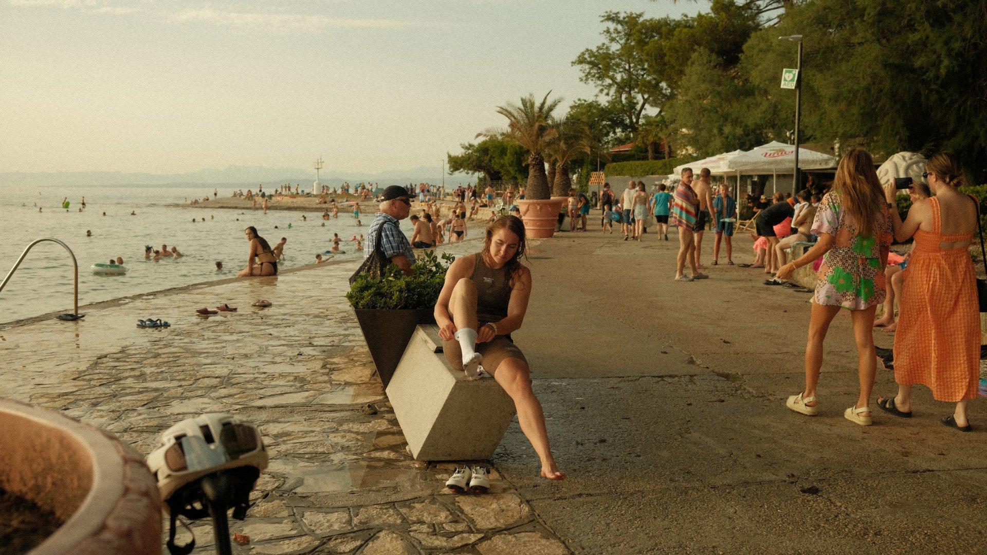 Model wearing dark cycling apparel sitting on concrete barrier at seaside promenade with beachgoers in background