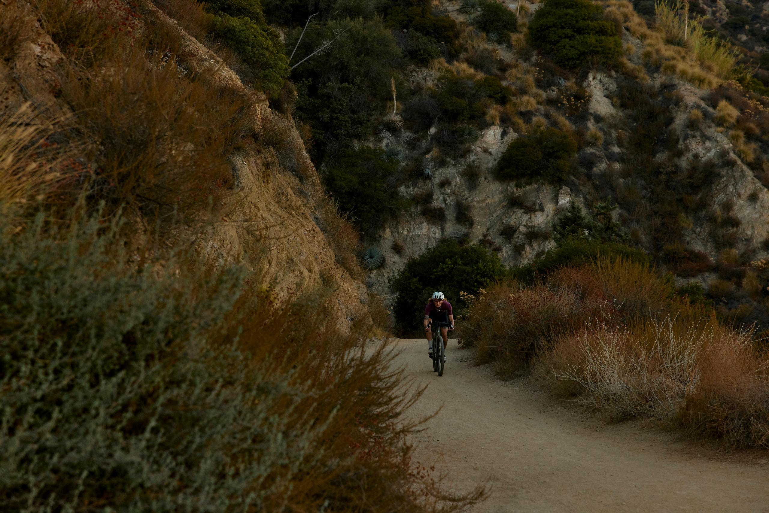 Cyclist in burgundy jersey riding through canyon path with rocky cliffs and vegetation