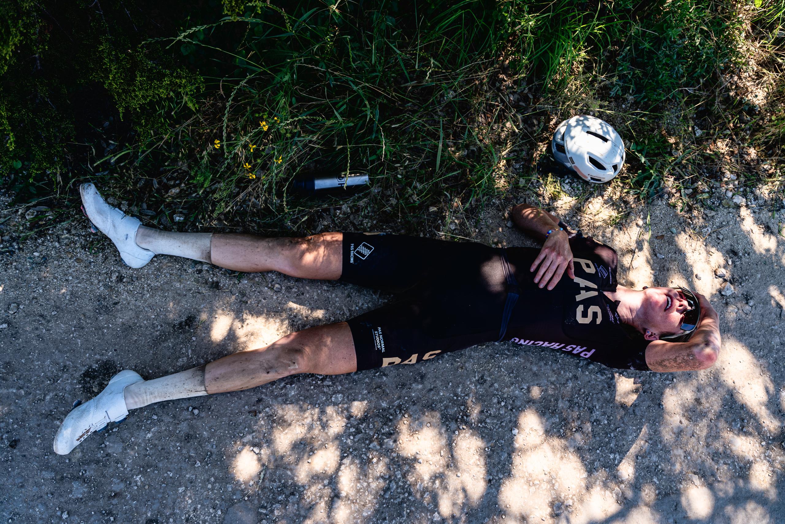 Model wearing black PAS collection jersey and bib shorts lying on asphalt road with cycling helmet nearby
