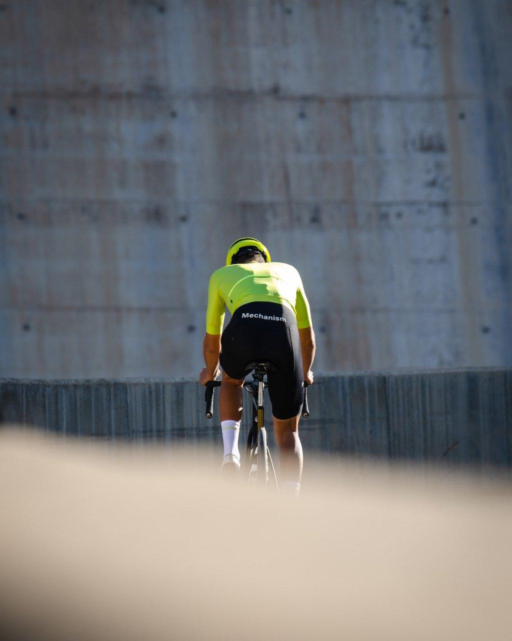 Model wearing green Mechanism jersey and black shorts in aerodynamic position on time trial bike