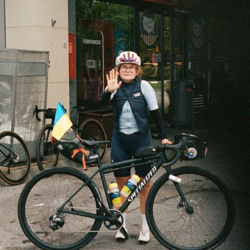 Model wearing navy vest over white base layer with white helmet, standing with bicycle outside storefront