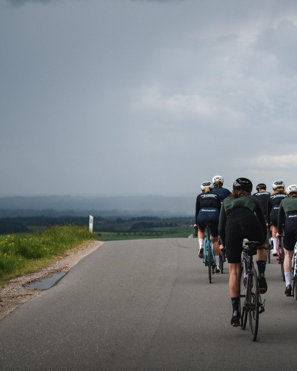 Group of cyclists wearing dark Mechanism jerseys riding on country road under cloudy sky