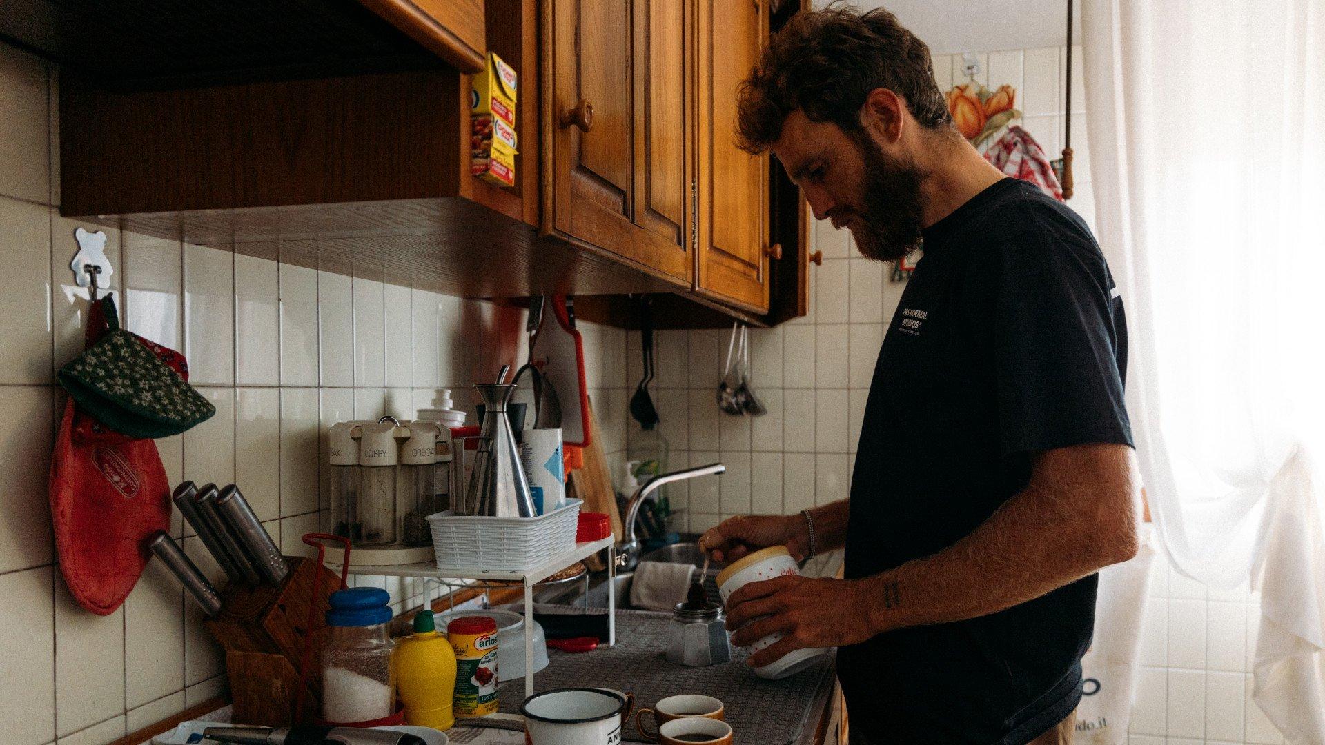 Model wearing black t-shirt with chest logo washing dishes in kitchen