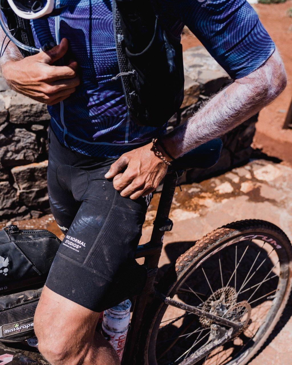 Model wearing blue patterned jersey and black shorts with bike in desert terrain
