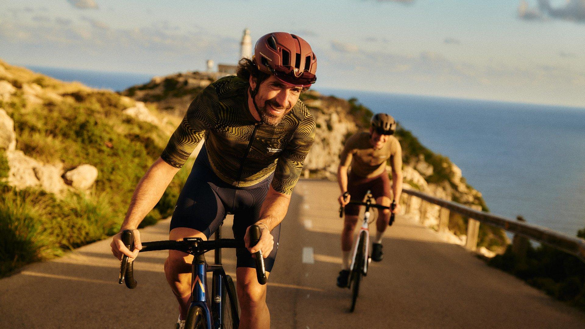 Model wearing brown patterned jersey and black shorts cycling on coastal road with second cyclist in background