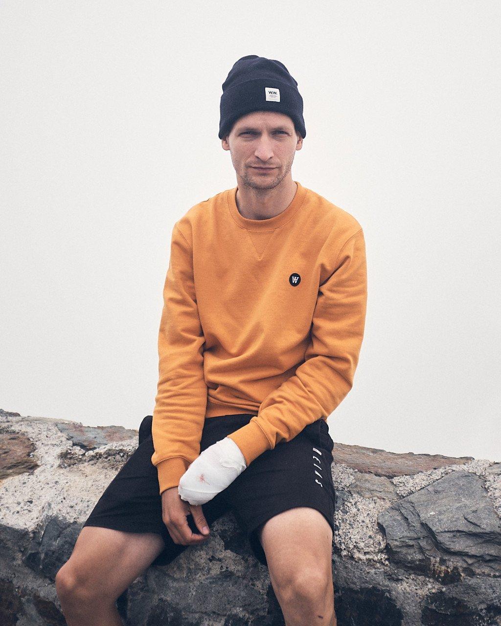 Model wearing orange sweatshirt with logo and navy beanie, sitting on rocks