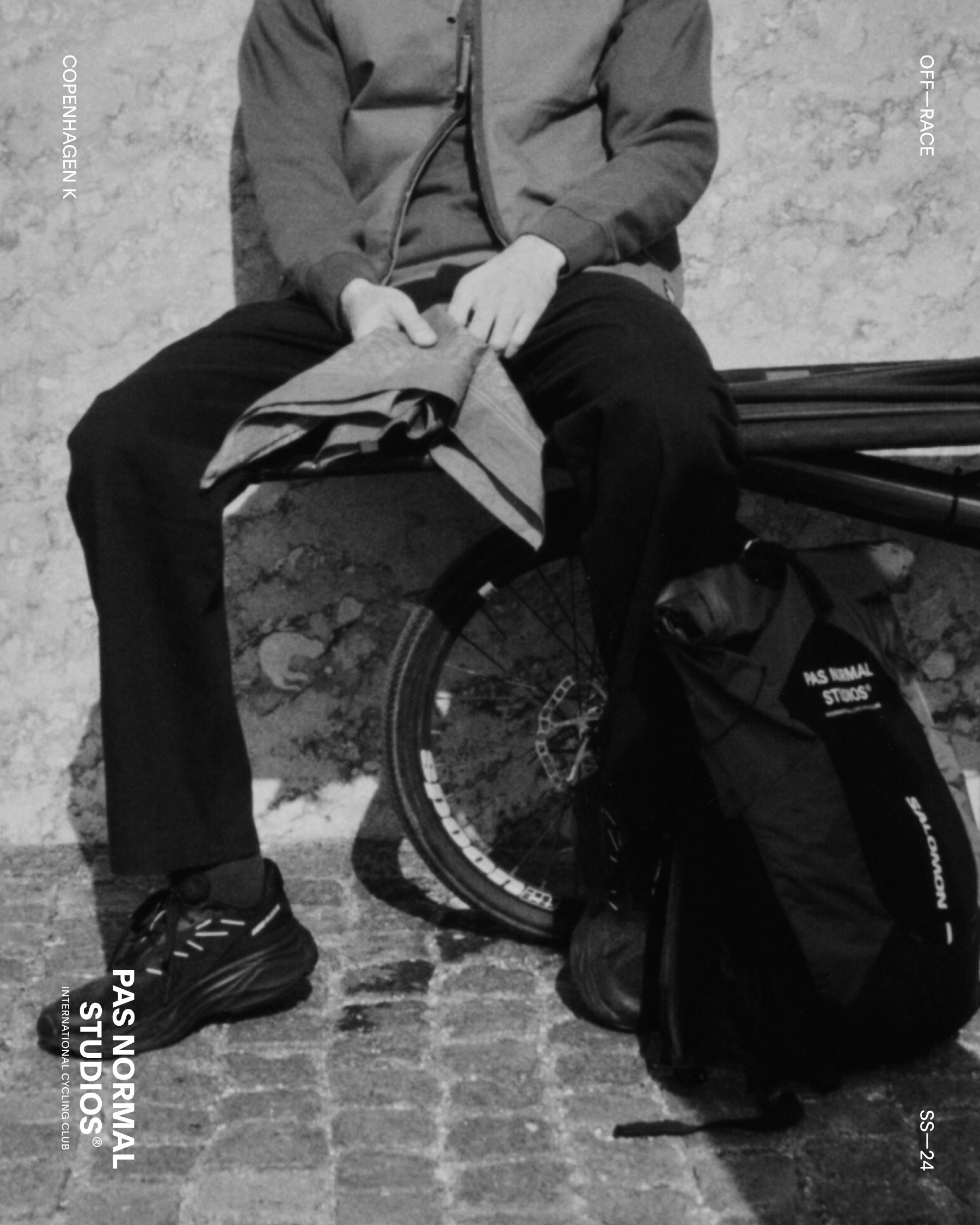 Model wearing grey hoodie and black pants sitting with bicycle and cycling gear in black and white photo