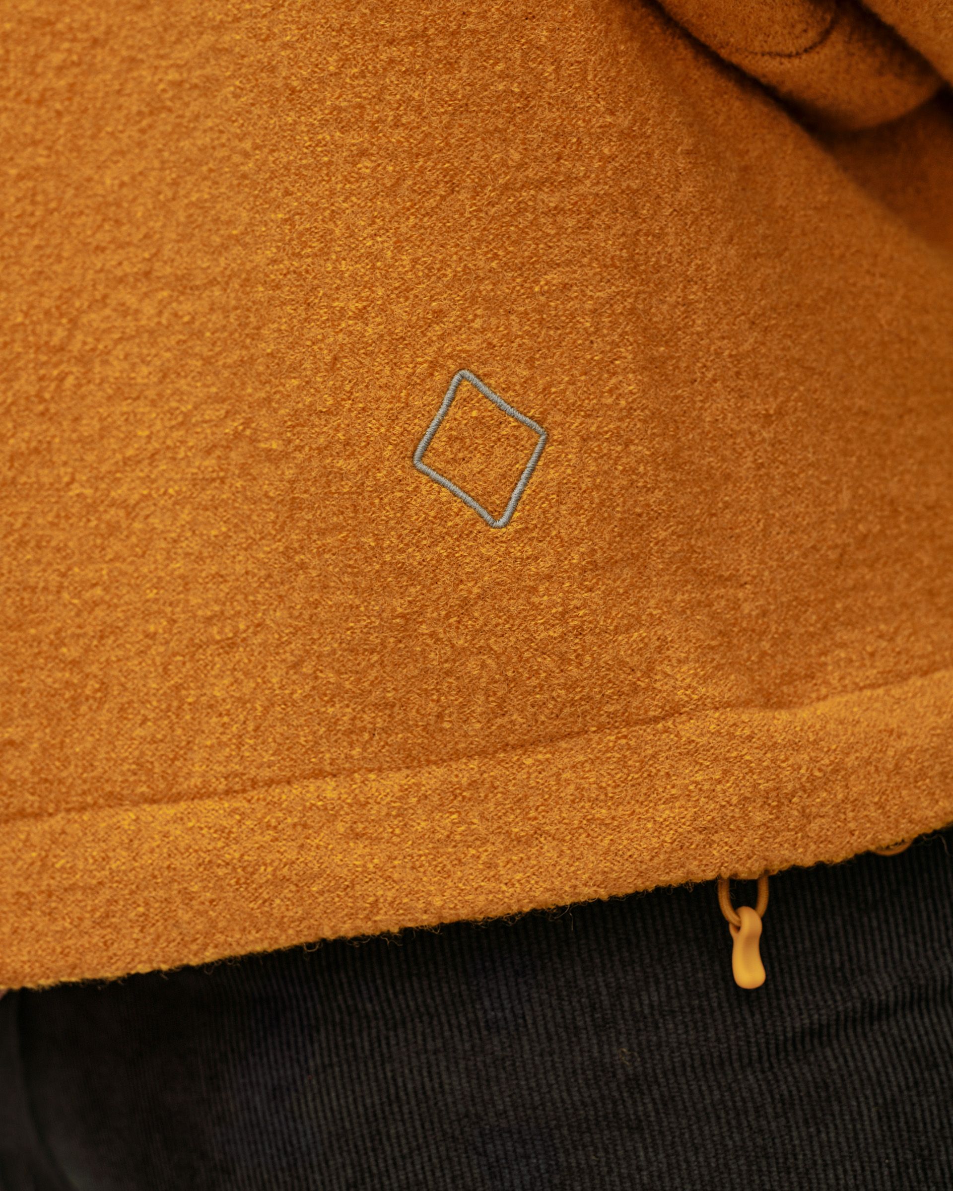 Ochre Off-Race Boiled Wool jacket, detail view of grey diamond logo embroidery and hem zip puller
