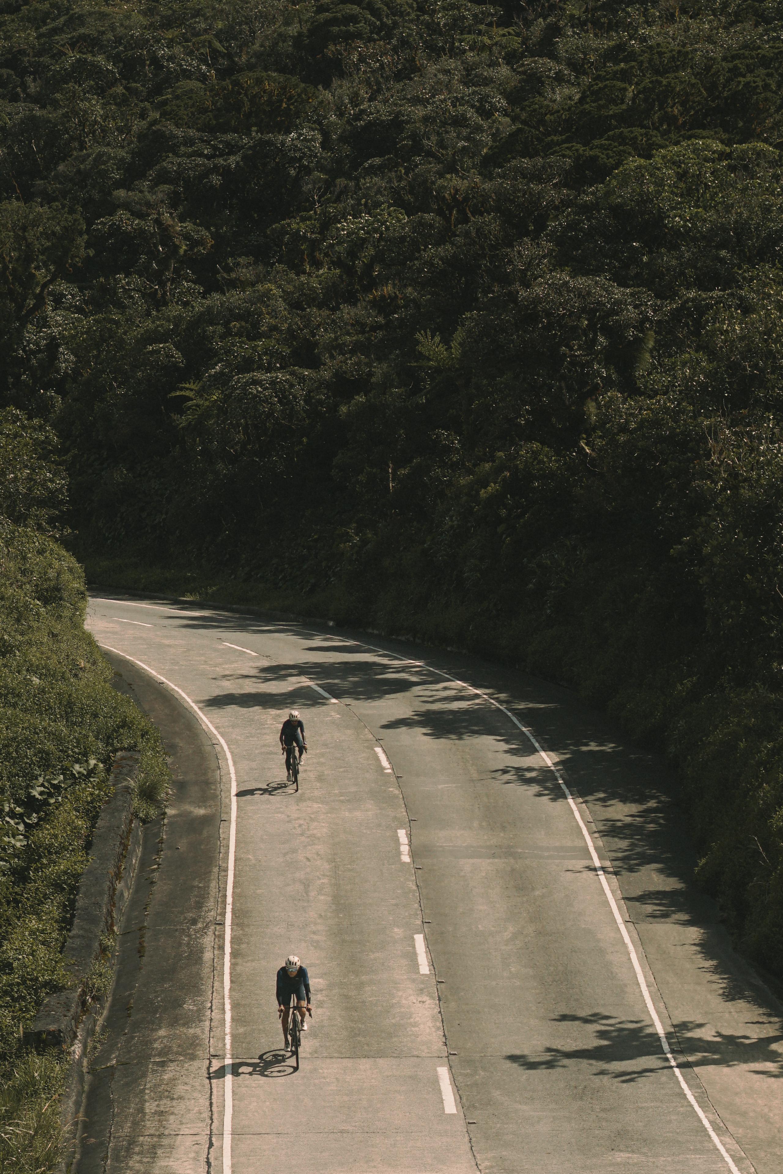 Two cyclists riding on winding road through forested area with dappled sunlight and shadows
