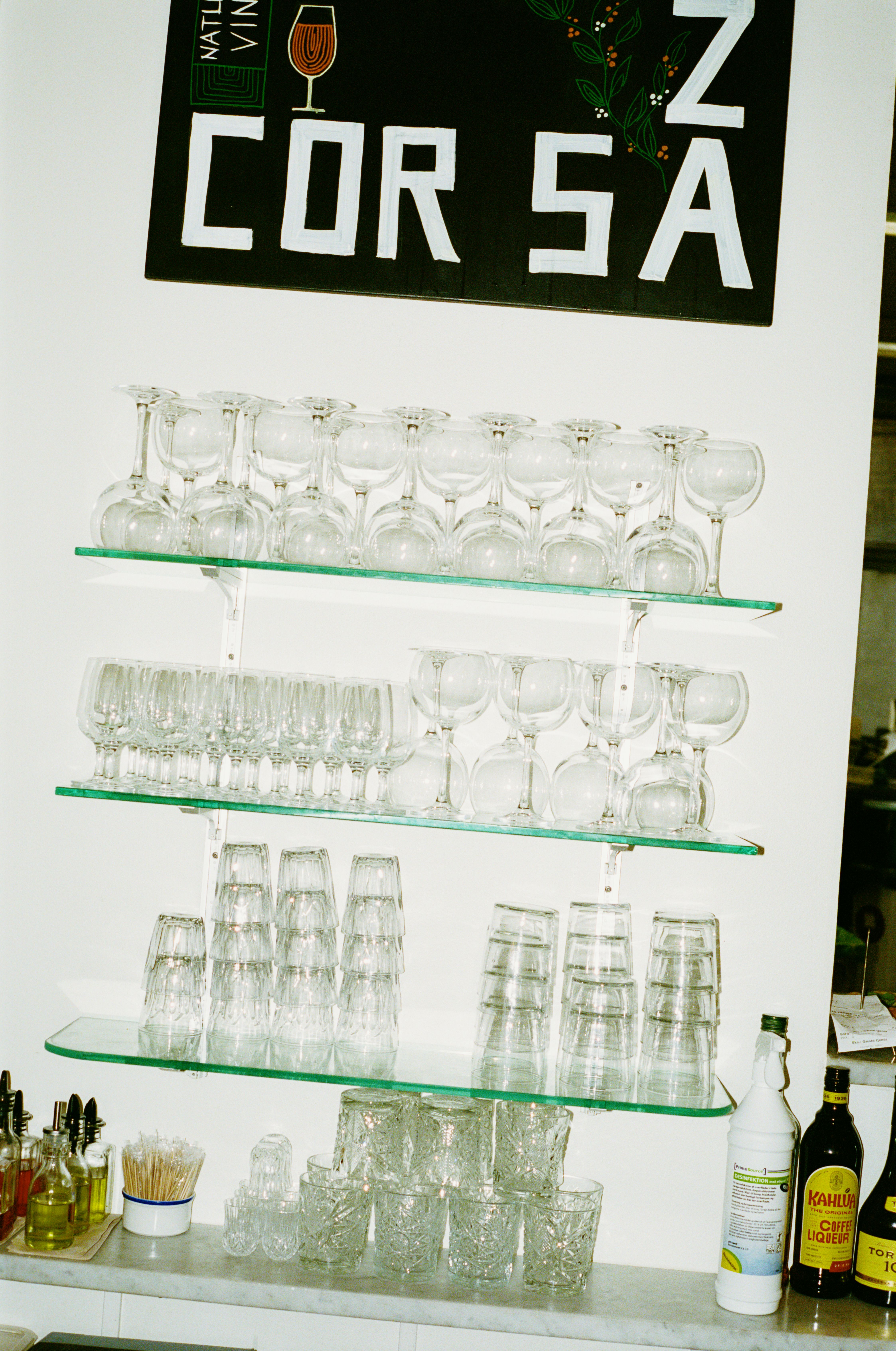 Glass shelving display with wine glasses and tumblers beneath Corsica poster