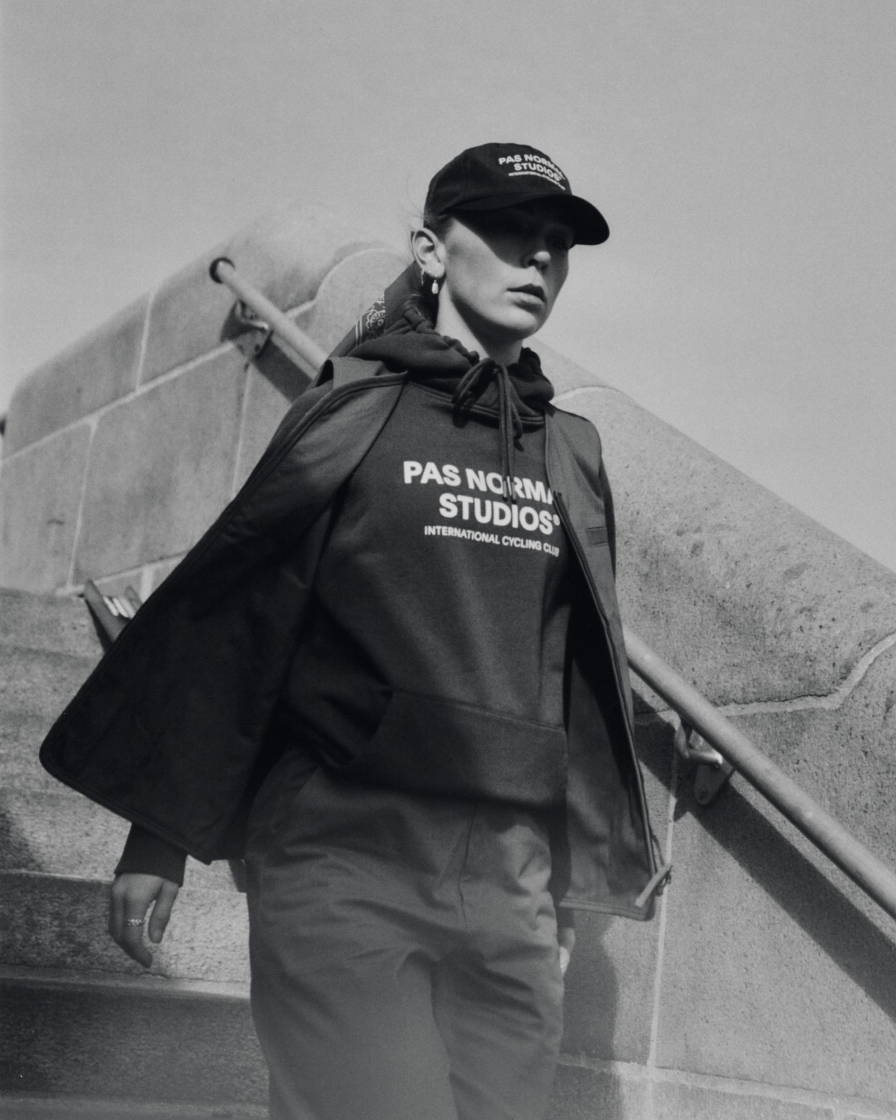 Model wearing dark hoodie with chest branding and black cap in urban concrete setting