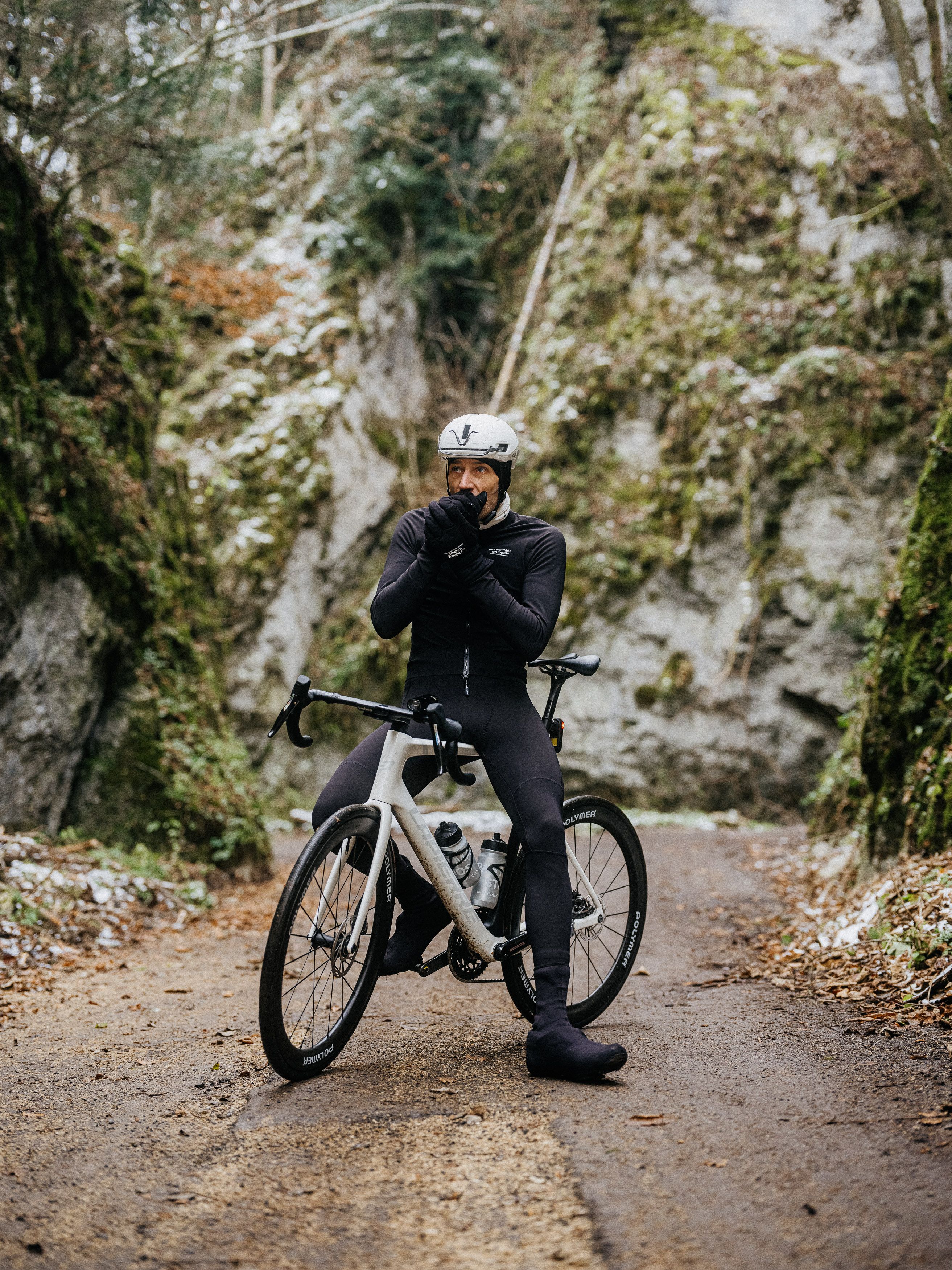 Model wearing black long-sleeve cycling kit with white helmet, standing beside white time trial bike on forest path