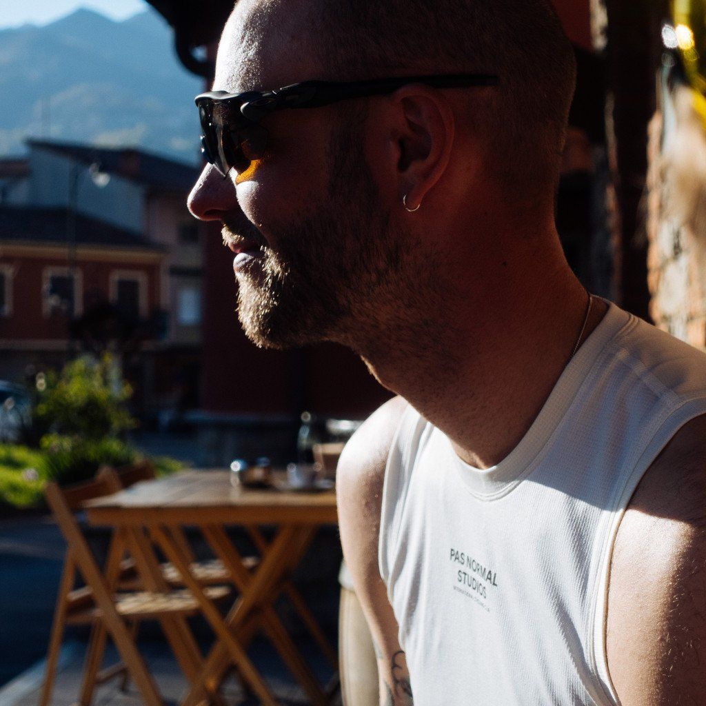 Model wearing white t-shirt with chest branding in profile, sunglasses, outdoor cafe setting
