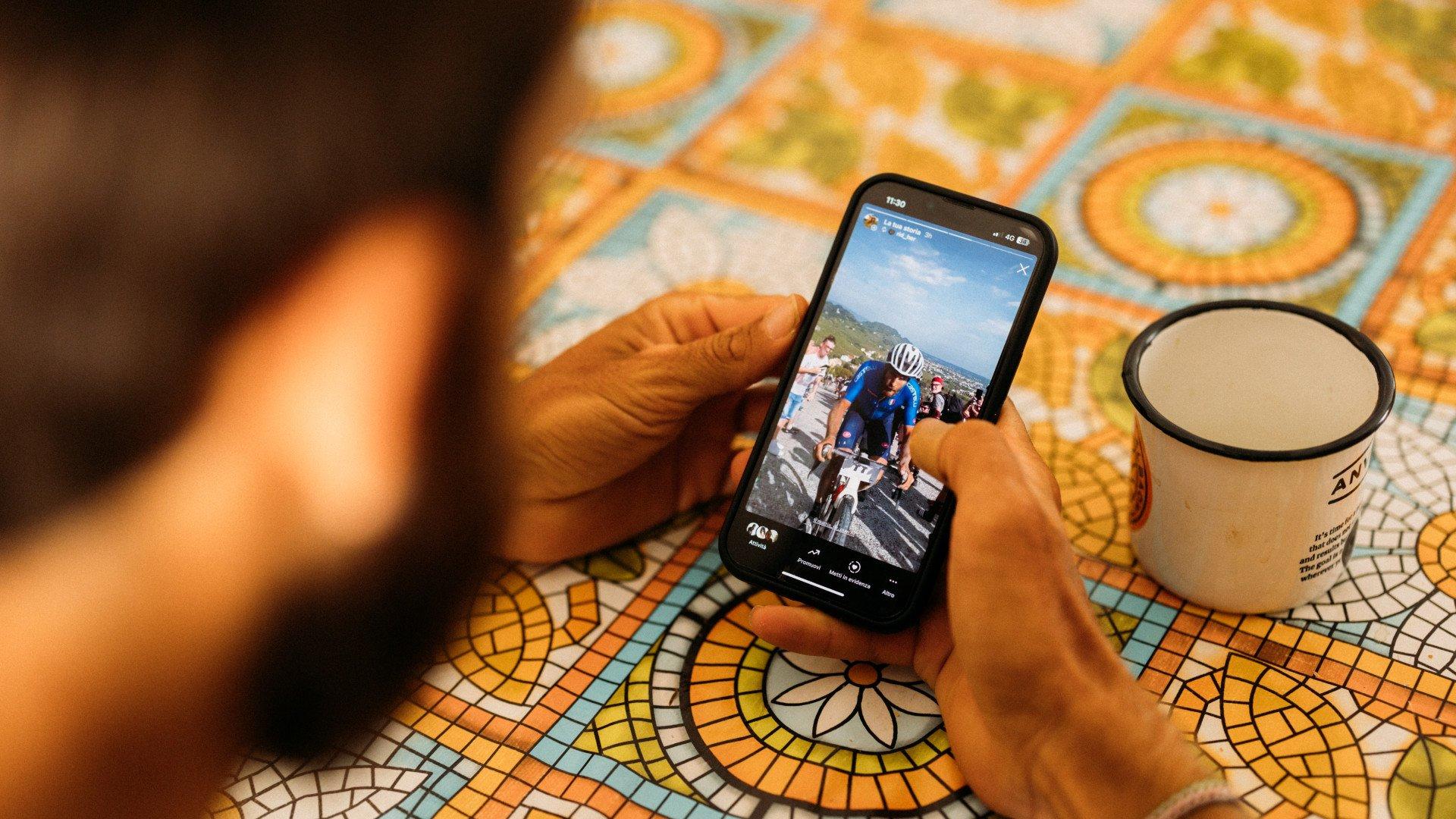 Person viewing cycling footage on smartphone while having coffee at patterned tile table