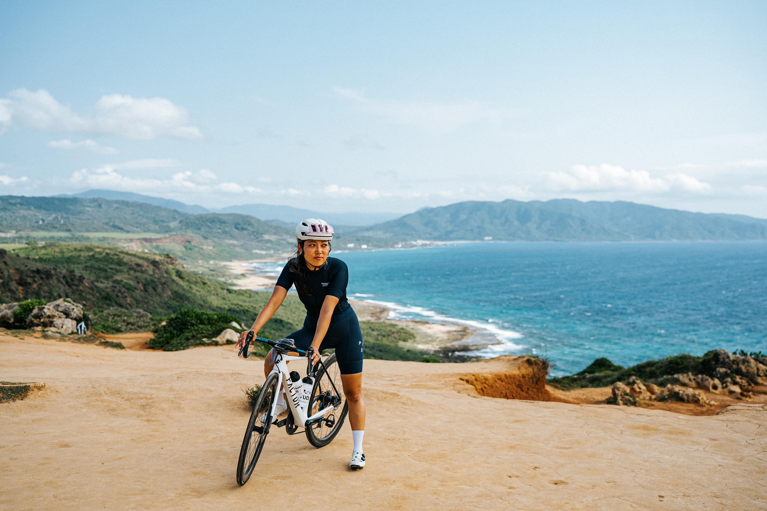 Model wearing Women's black cycling jersey and shorts with white helmet, walking bike on coastal dirt road with ocean view