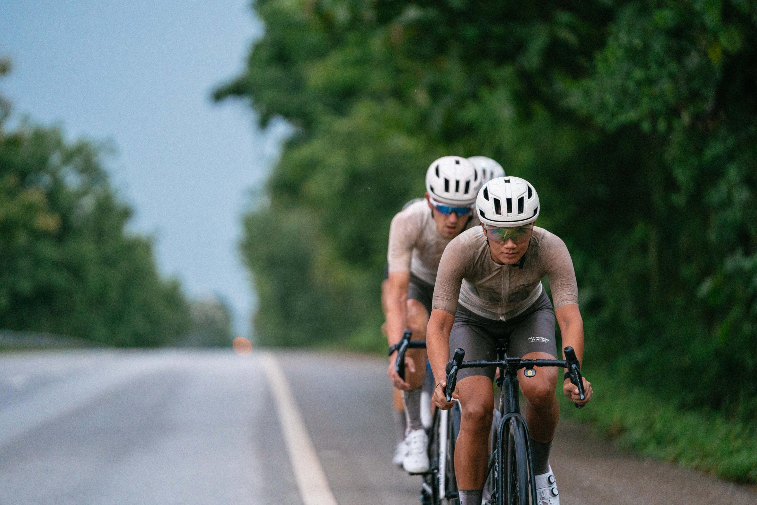 Two cyclists in aerodynamic position on time trial bikes riding on rural road with green foliage