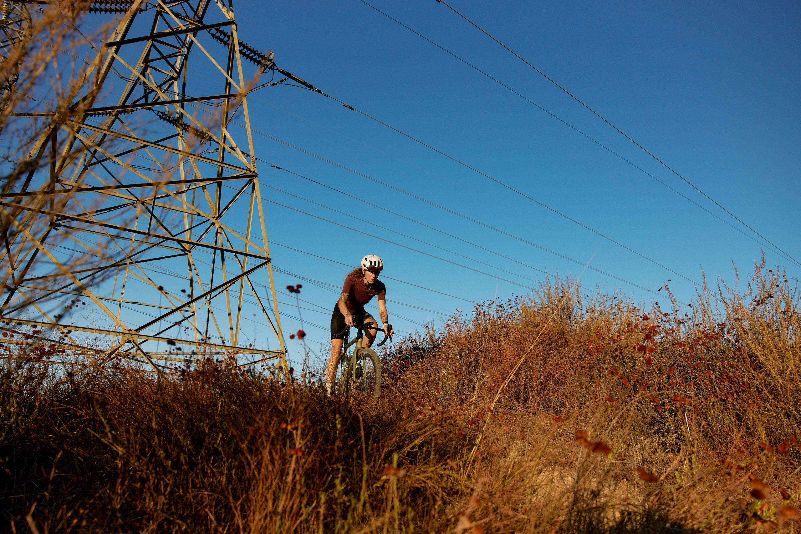 Model wearing brown jersey and black shorts cycling on hillside trail beneath power lines