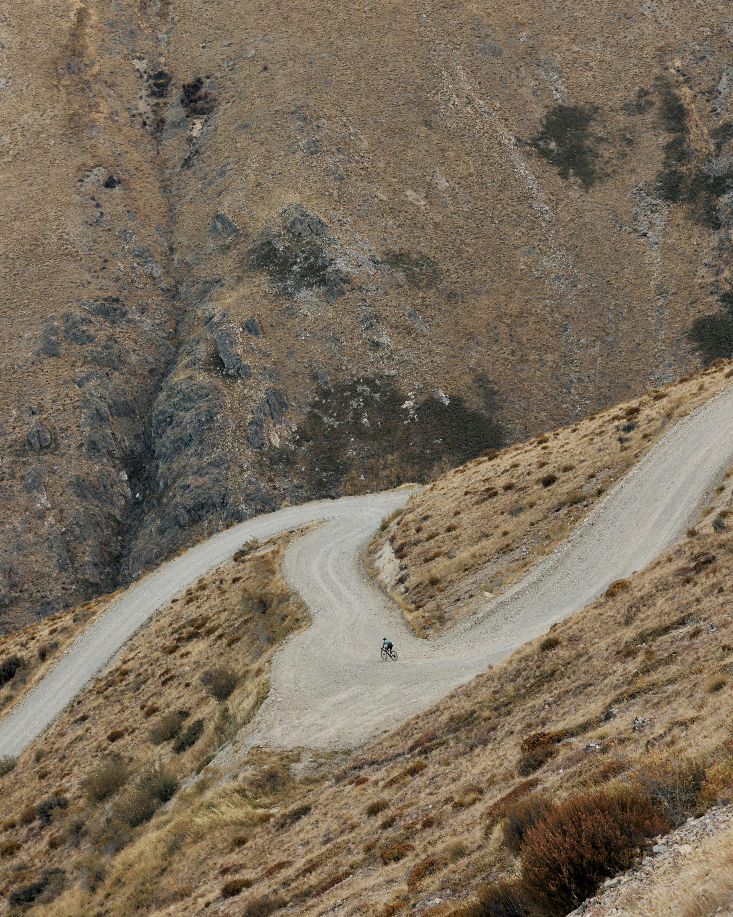 Cyclist on winding mountain road through arid landscape with golden grass and rocky terrain