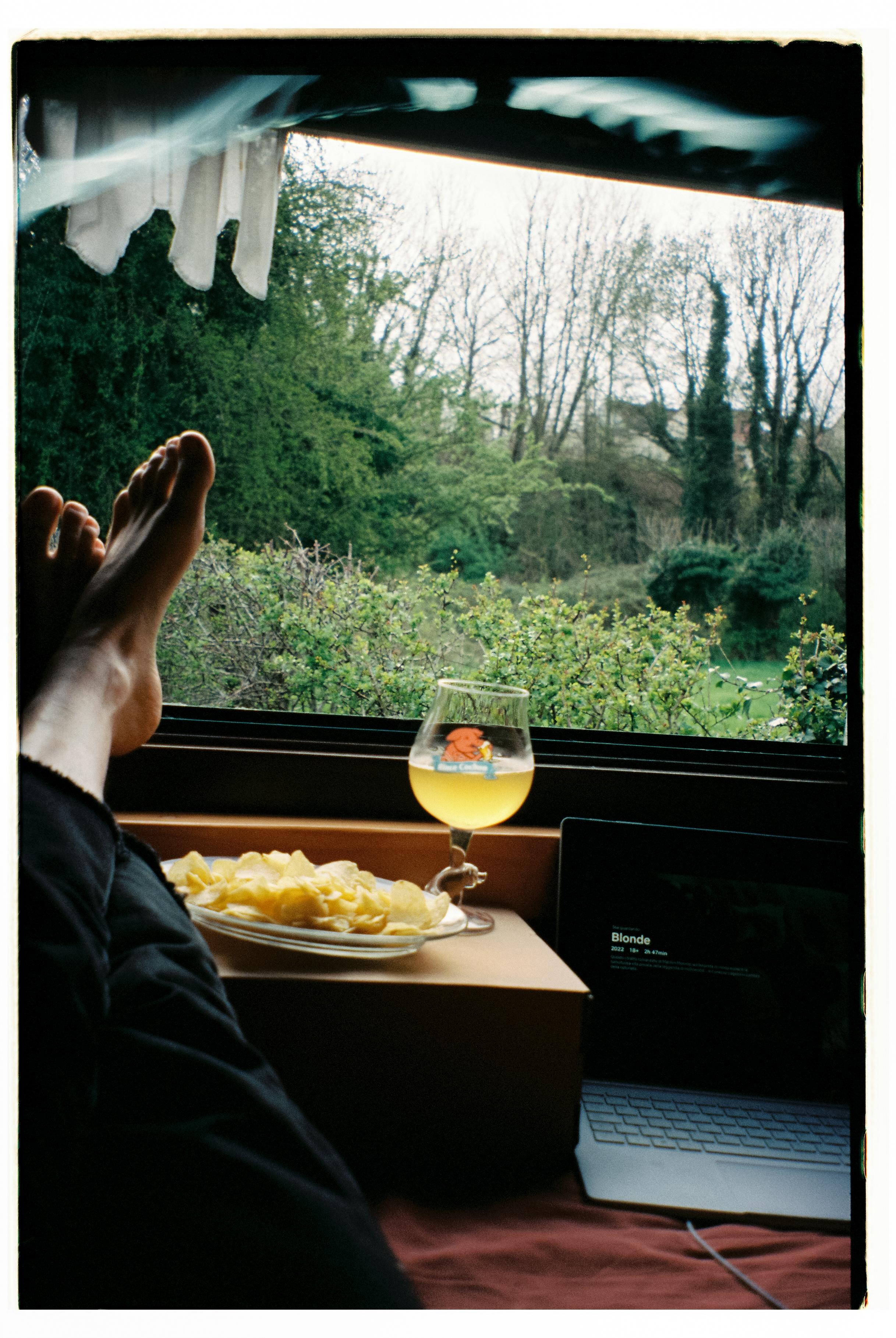 Person relaxing indoors with feet up, viewing garden through window with laptop, drink and snacks nearby
