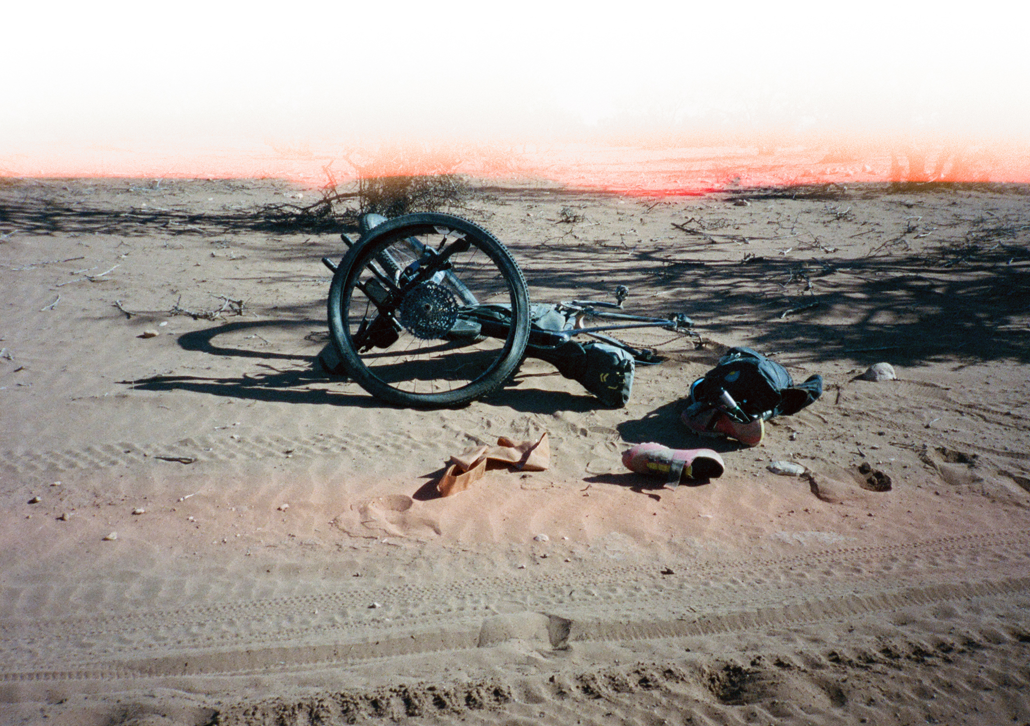 Cyclist resting on sandy ground beside bicycle wheel in desert landscape with orange horizon