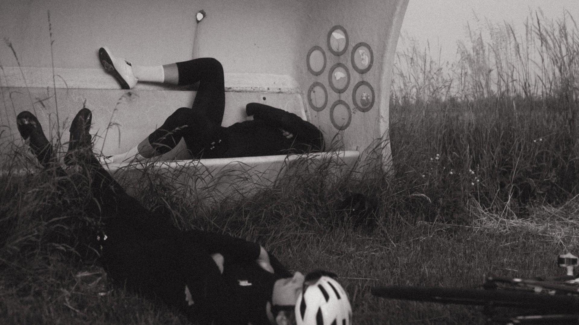Cyclist in black clothing resting on concrete ledge with bicycle helmet visible in foreground