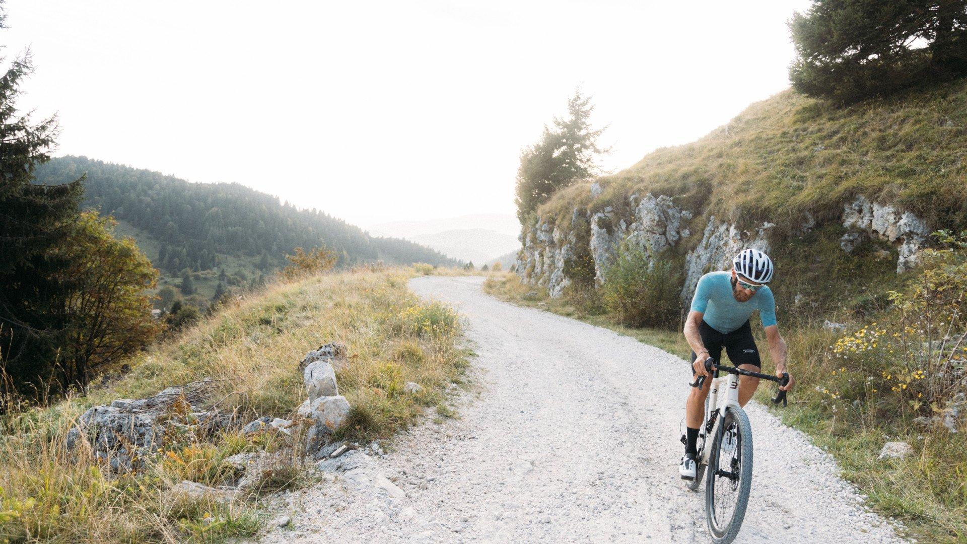 Model wearing light blue jersey cycling on gravel road through mountainous terrain