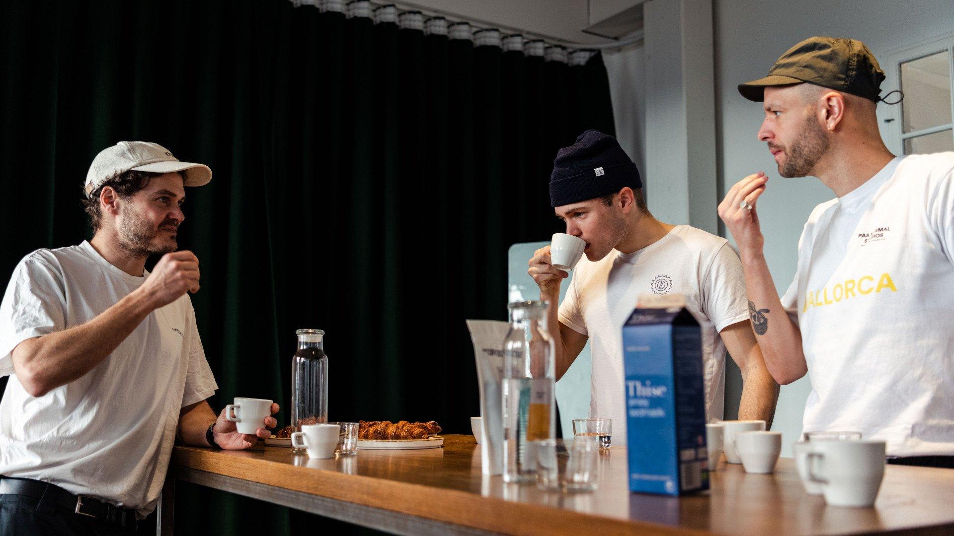 Three men wearing casual apparel including caps and t-shirts having coffee at wooden table in modern interior
