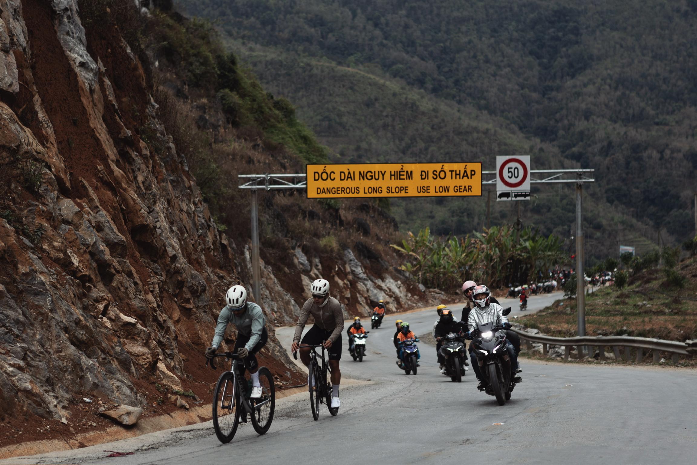 Two cyclists riding time trial bikes on mountain road with motorcycles and warning signs in Vietnam