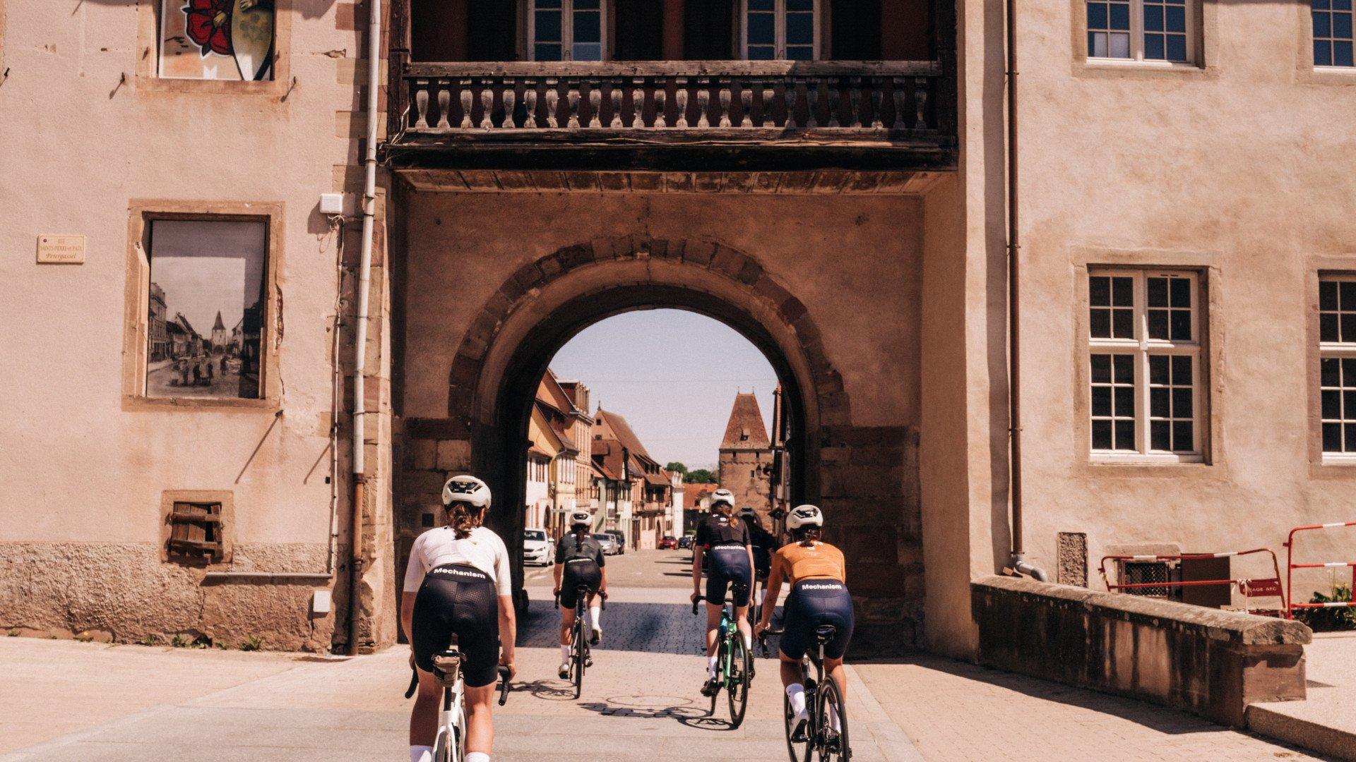 Cyclists riding through historic stone archway in European town wearing cycling apparel