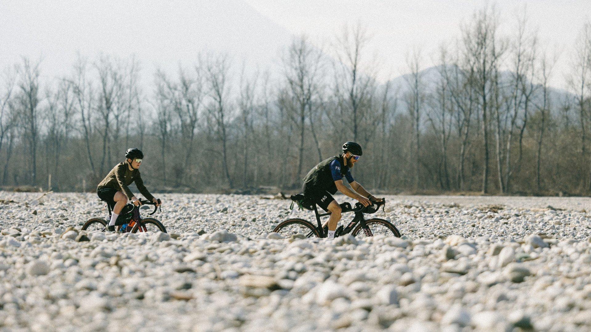 Two cyclists riding gravel bikes across rocky terrain with bare trees in background
